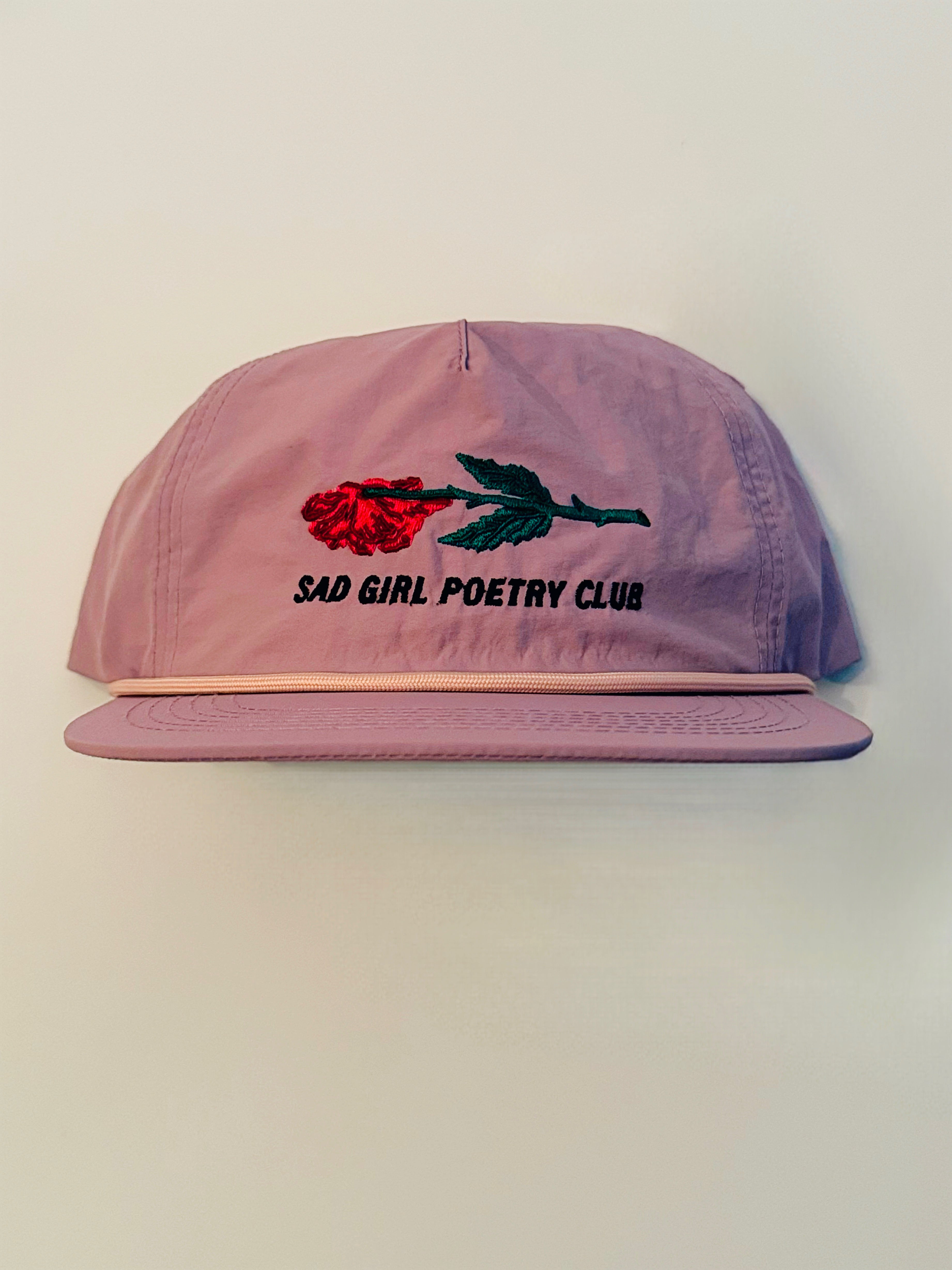 "Embroidered lavender hat featuring a red flattened rose with a green stem, and black text underneath that reads 'Sad Girl Poetry Club'"