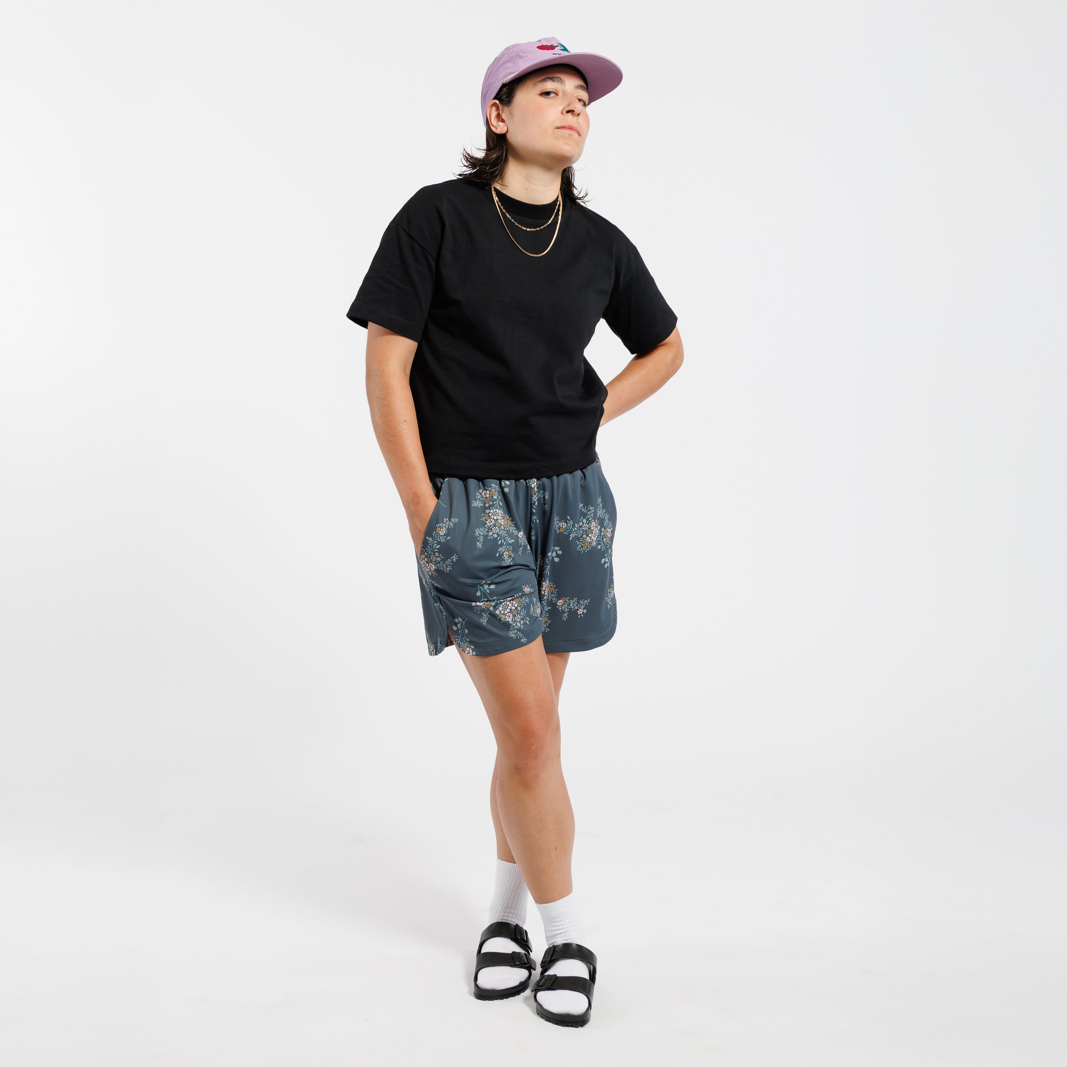 Person wearing a black t-shirt, Floral-patterned shorts, and a pink cap on a white background