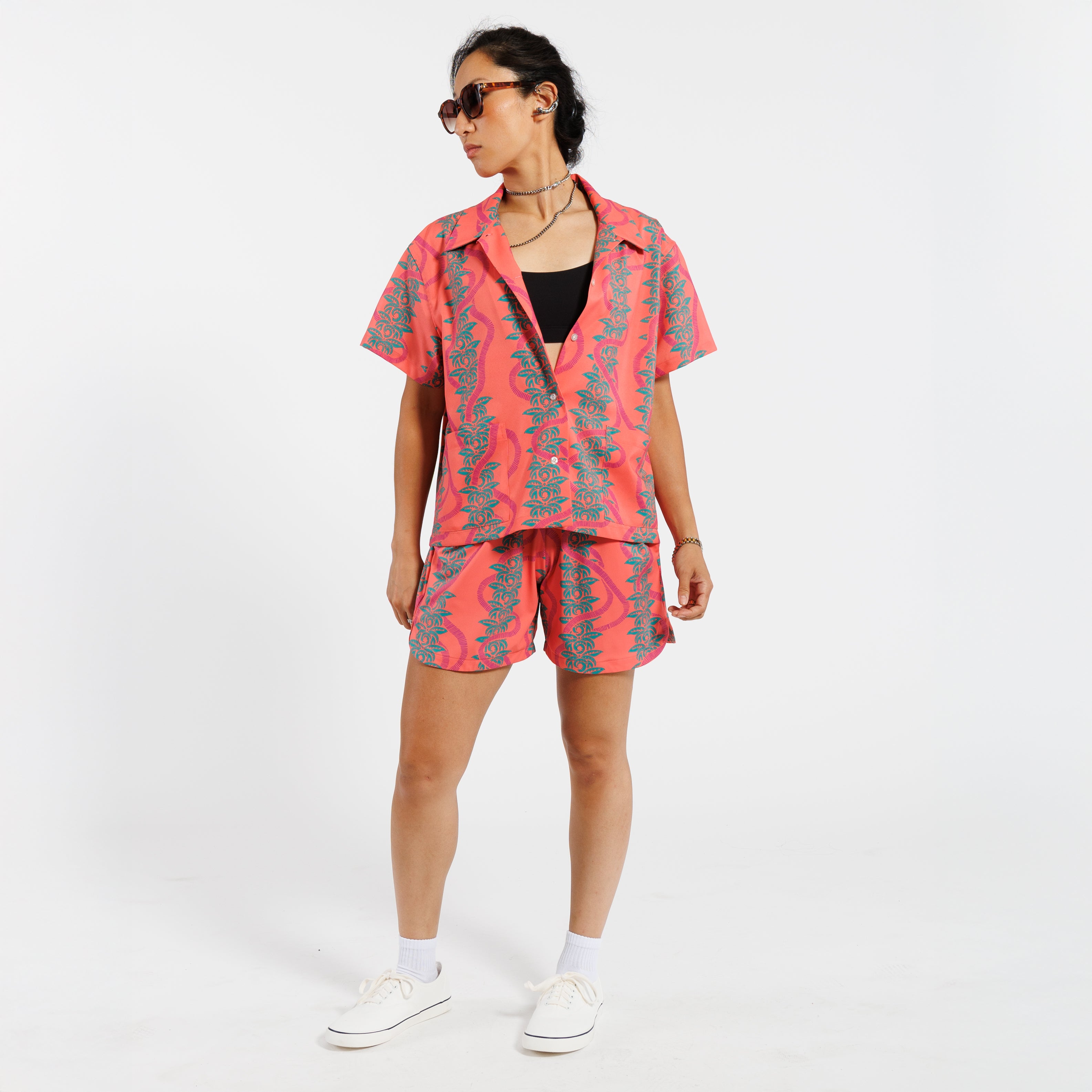Woman wearing a pink button-down shirt with green leaf pattern, paired with matching set shorts and a white undershirt, standing against a white background.