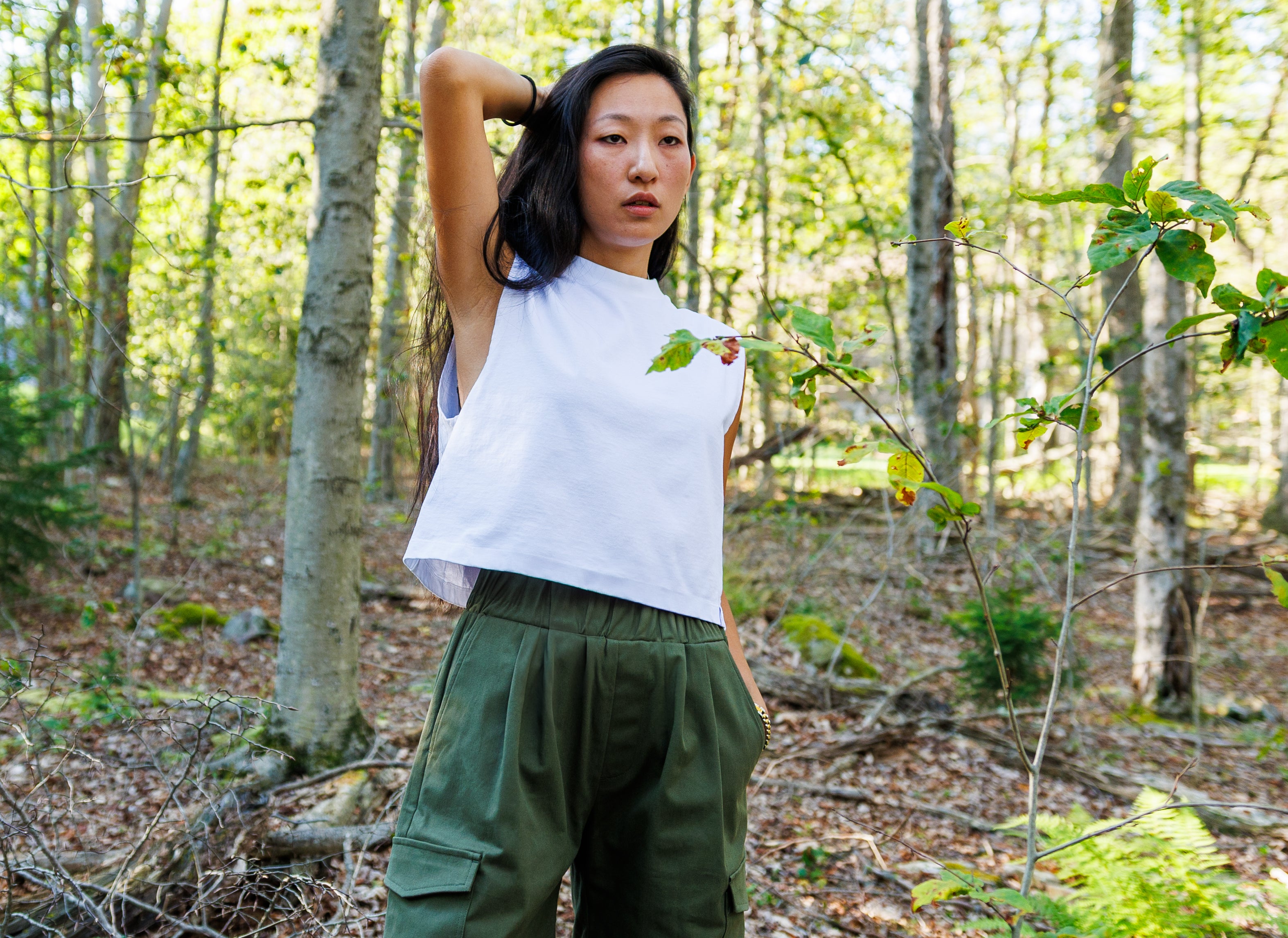 Woman standing in a forest wearing a white top and green pants.