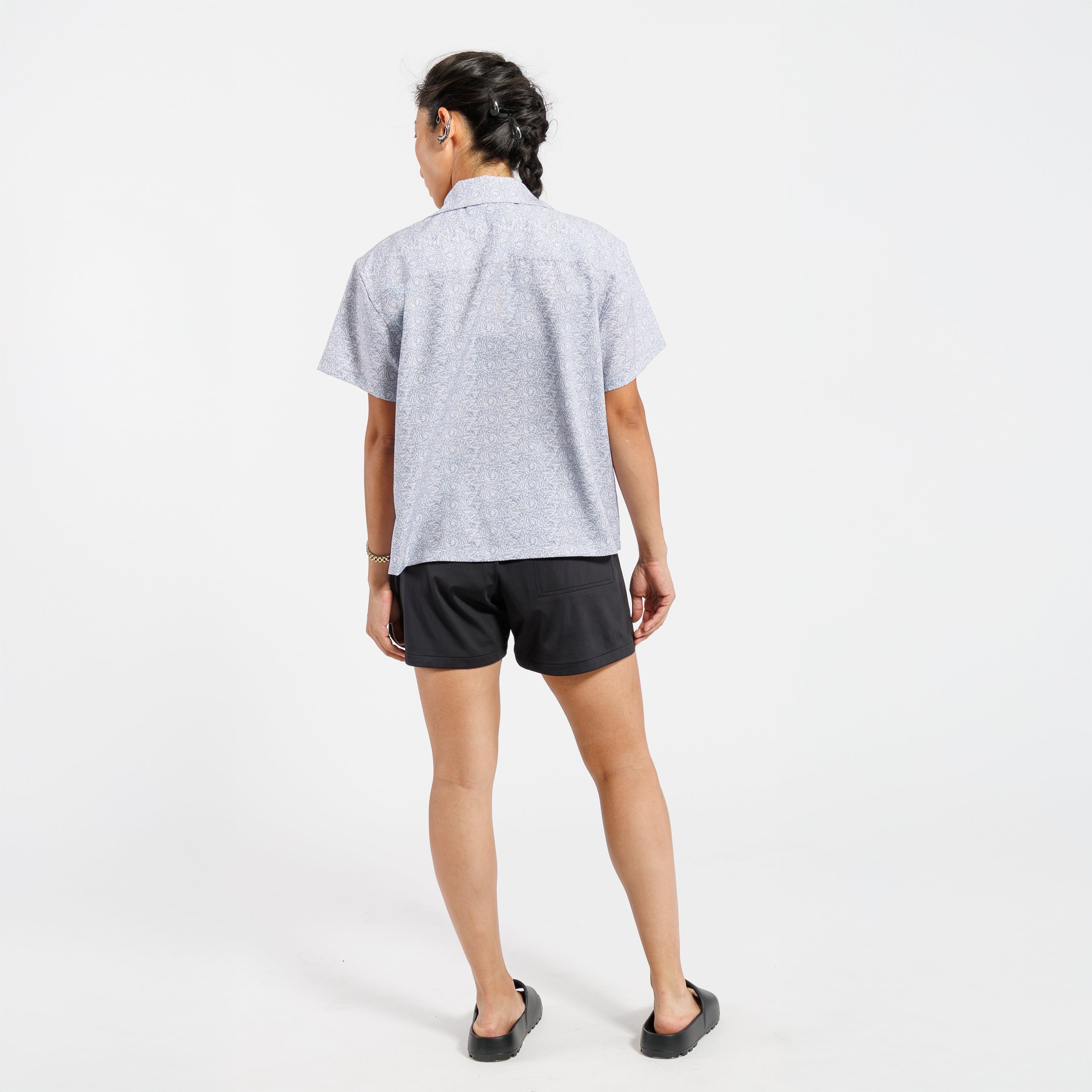 Back view of a woman wearing a short-sleeve button-down shirt with a subtle white leaf pattern, standing against a white background.