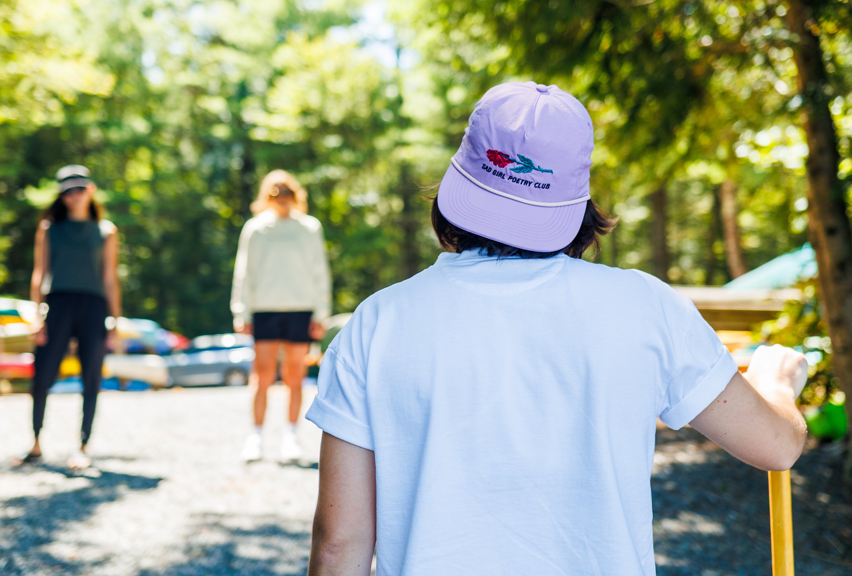 Person wearing a purple cap with a logo, standing outdoors with trees and people in the background.