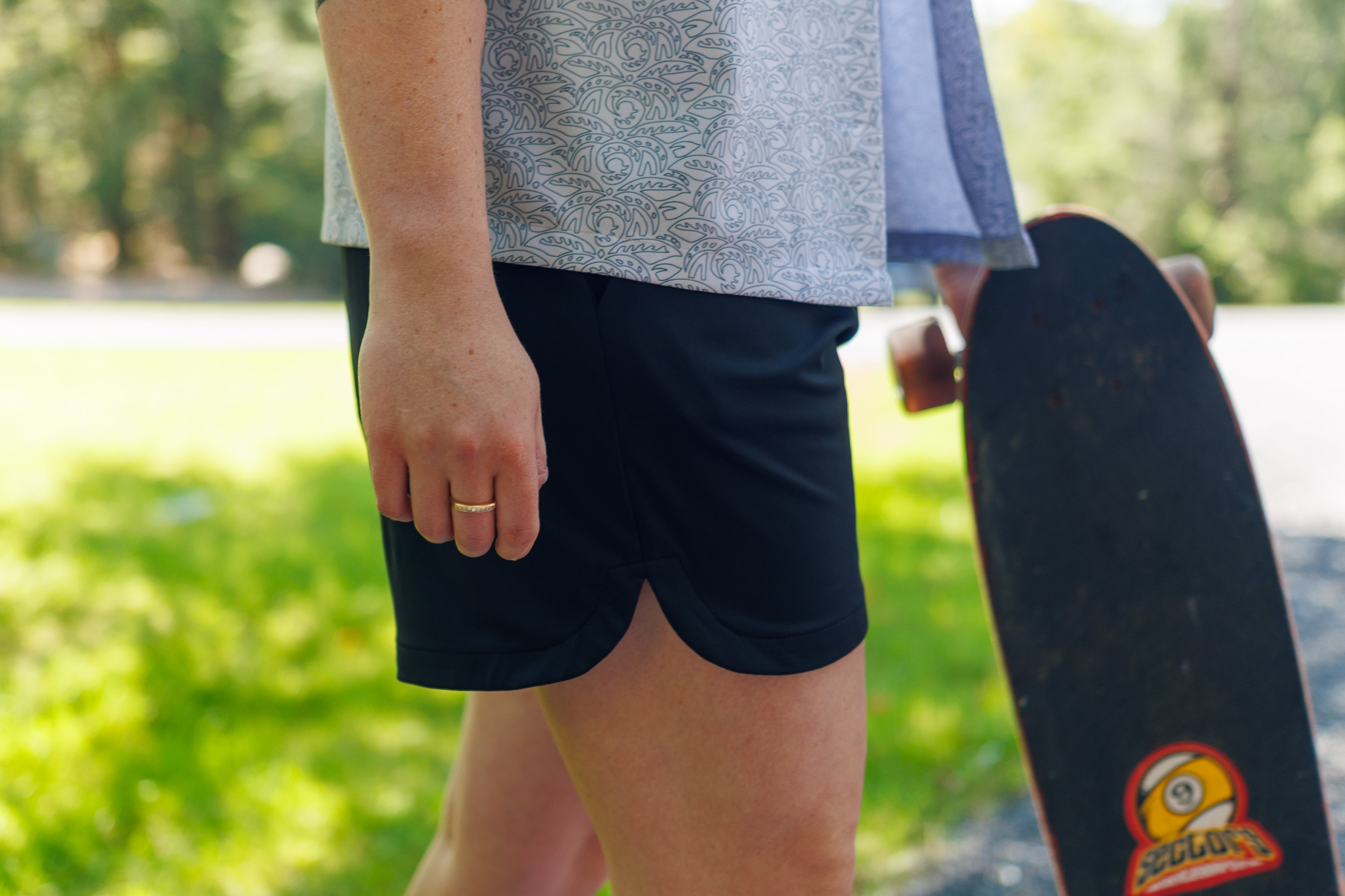 Side view of a person wearing black shorts and a white leaf-patterned shirt, holding a skateboard in one hand.