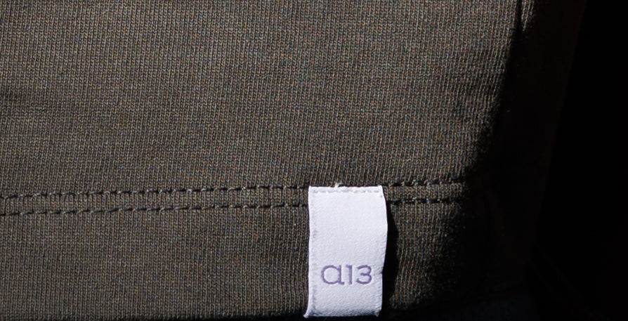 White logo label 'a13' in stitched on gray fabric against jean pant.