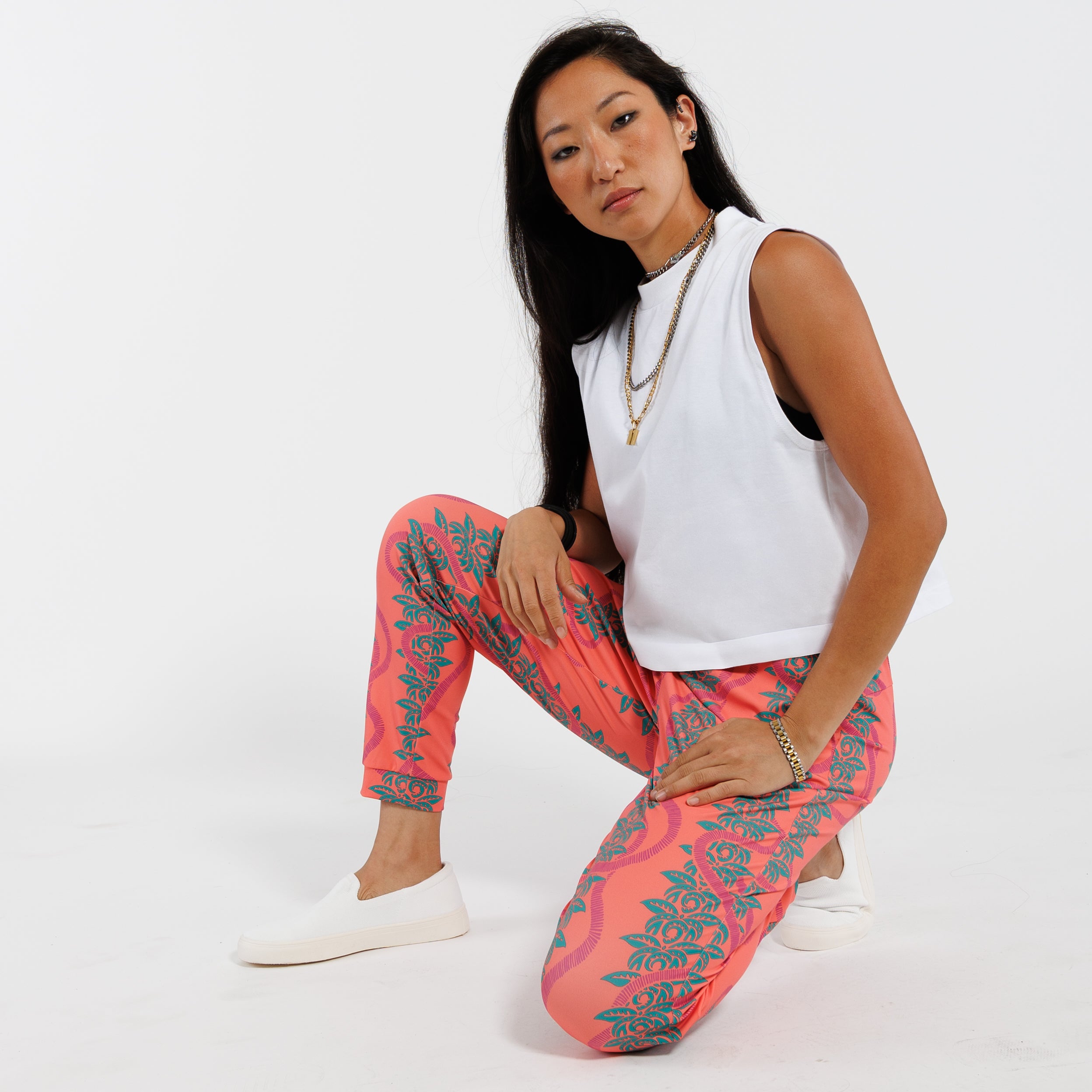 Woman bent on left knee wearing a white cropped tank top and pink patterned with green leaves pants on a white background