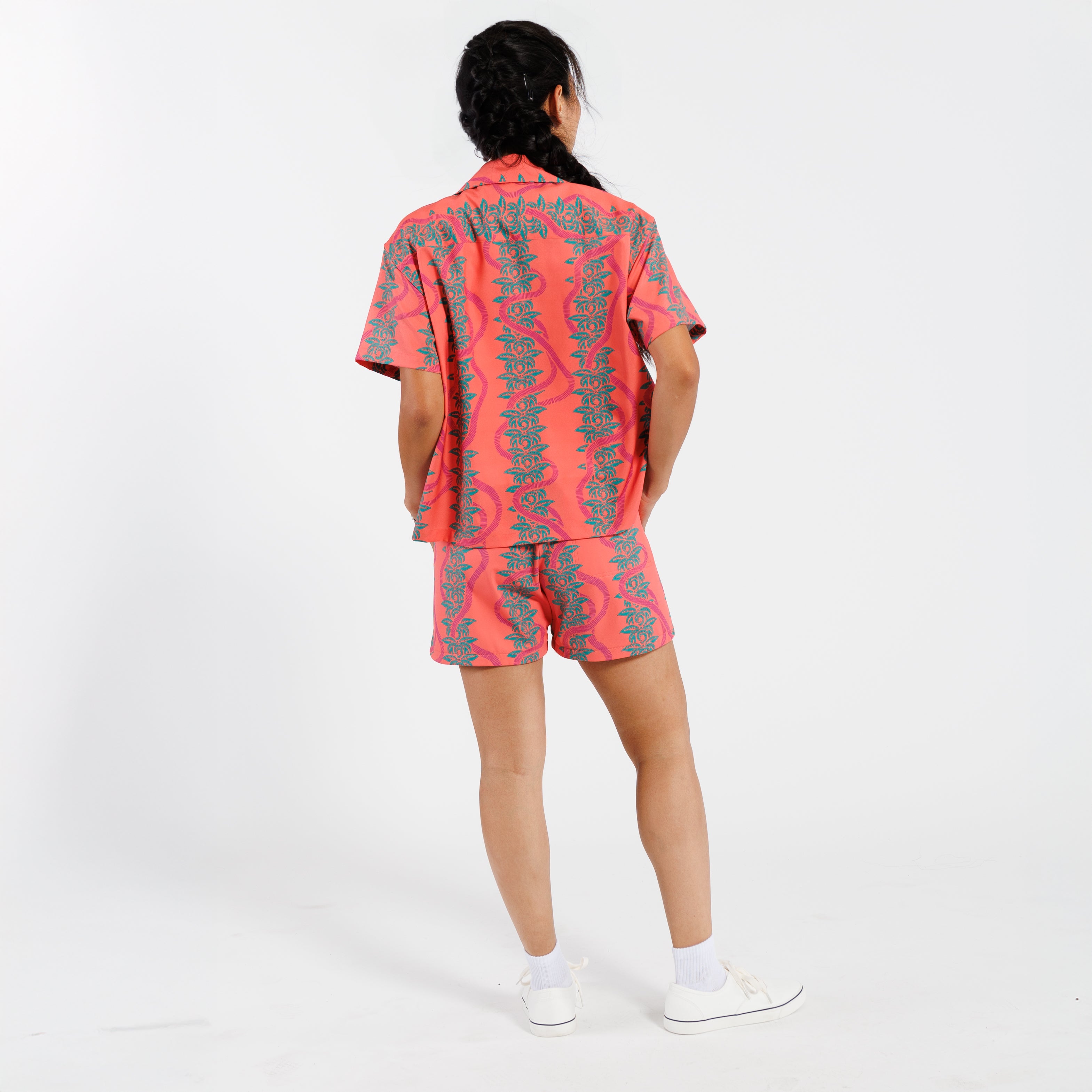 Back view of a woman wearing a pink shirt with a green leaf pattern, paired with matching shorts, standing against a white background.