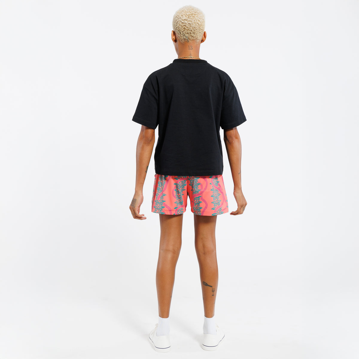 Back view of a person wearing a black t-shirt and pink shorts with a green leaf pattern, standing in front of a white background