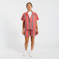 Woman wearing a pink button-down shirt with a green leaf pattern, paired with matching shorts and a white undershirt, standing against a white background.