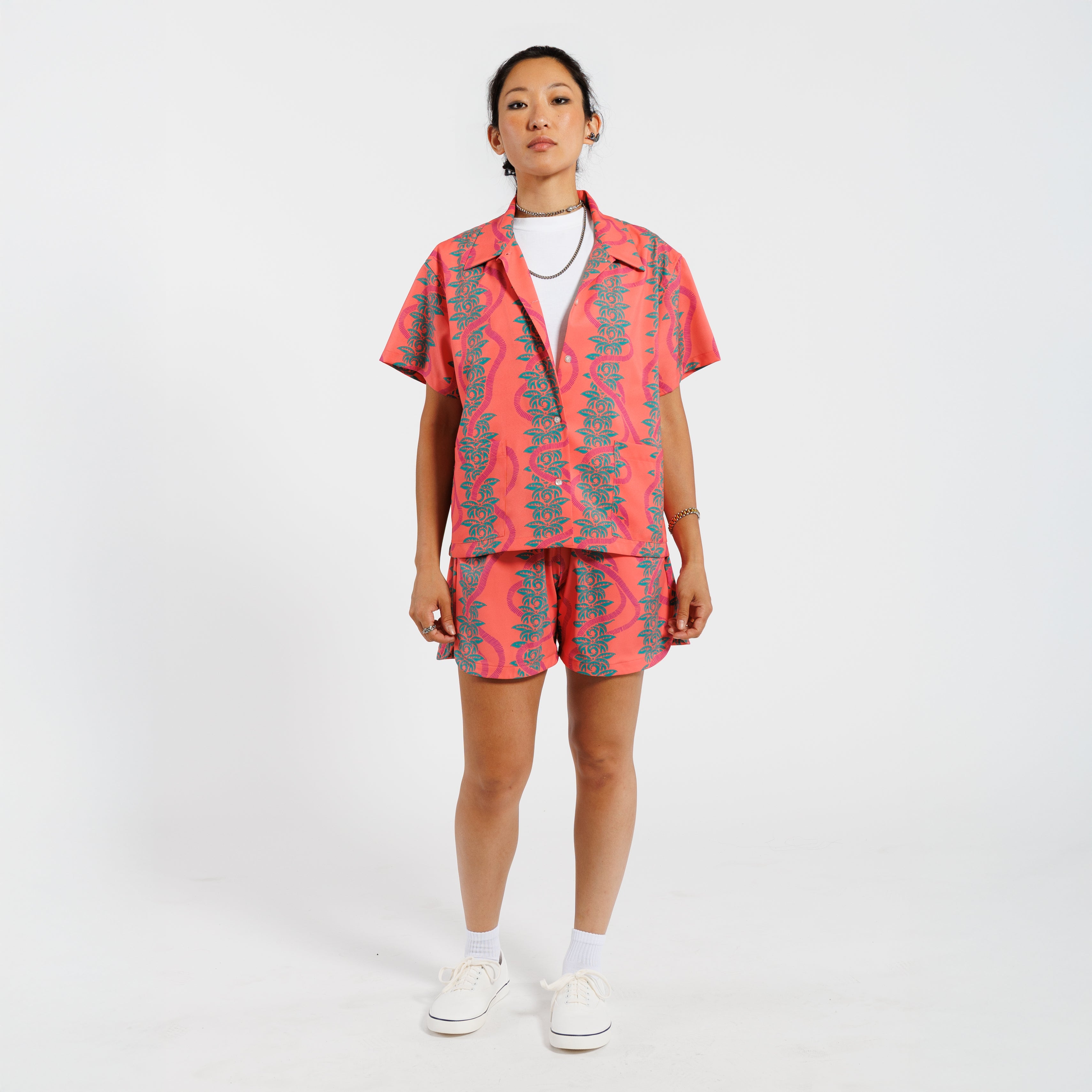 Woman wearing a pink button-down shirt with a green leaf pattern, paired with matching shorts and a white undershirt, standing against a white background.