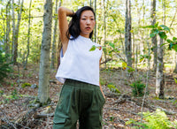 Woman standing in a forest wearing a white top and green pants.