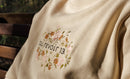 Cream sweatshirt with floral embroidery and text on a blurred natural background