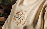 Cream sweatshirt with floral embroidery and text on a blurred natural background