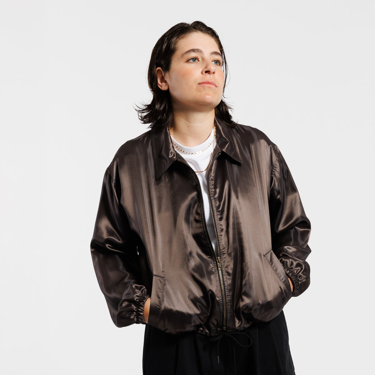 Person wearing a iridescent copper jacket with double zippers and black drawstrings, with both hands in front pocket, displayed on a white background.