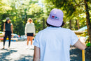 Person wearing a purple cap with a logo, standing outdoors with trees and people in the background.