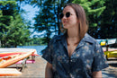 Woman wearing a floral button-down shirt standing in a forested boatyard.