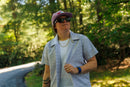 Person wearing sunglasses, a hat, and a white leaf-patterned button-down shirt over a white shirt, standing in a forest.