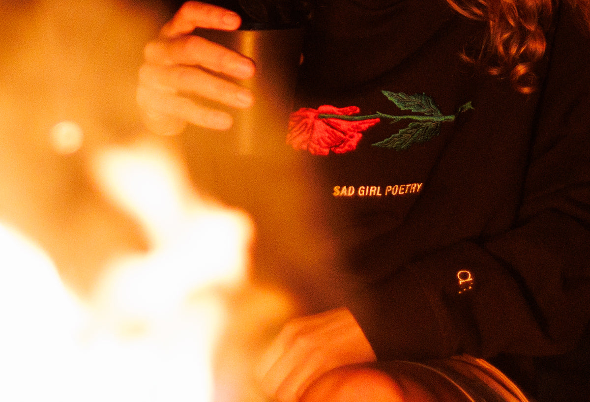 Woman with curly hair wearing a black hoodie in front of a bonfire, featuring a withered pink and green flower and white “Sad Girl Poetry” text on the chest, with an A13 logo on the sleeve.