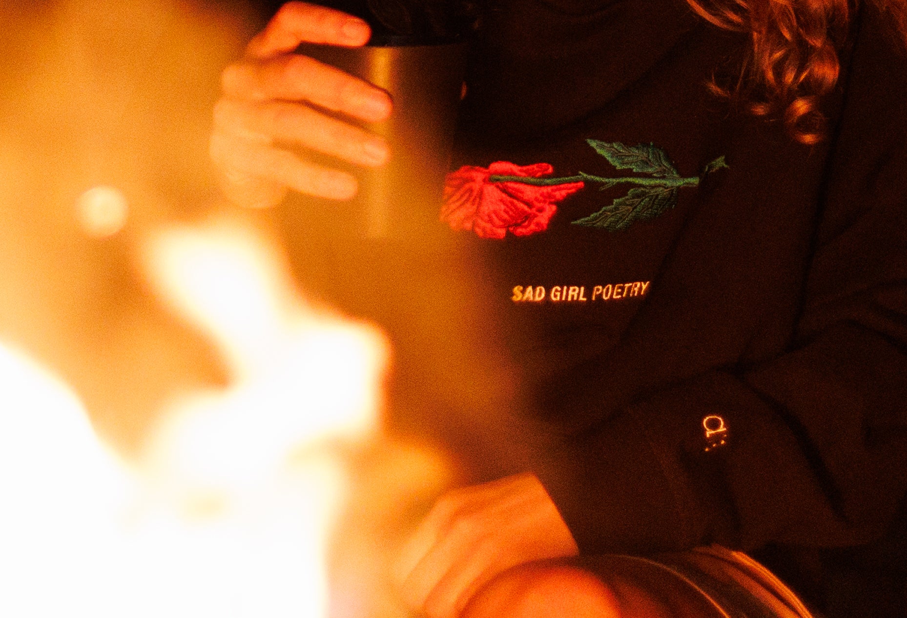 Woman with curly hair wearing a black hoodie in front of a bonfire, featuring a withered pink and green flower and white “Sad Girl Poetry” text on the chest, with an A13 logo on the sleeve.
