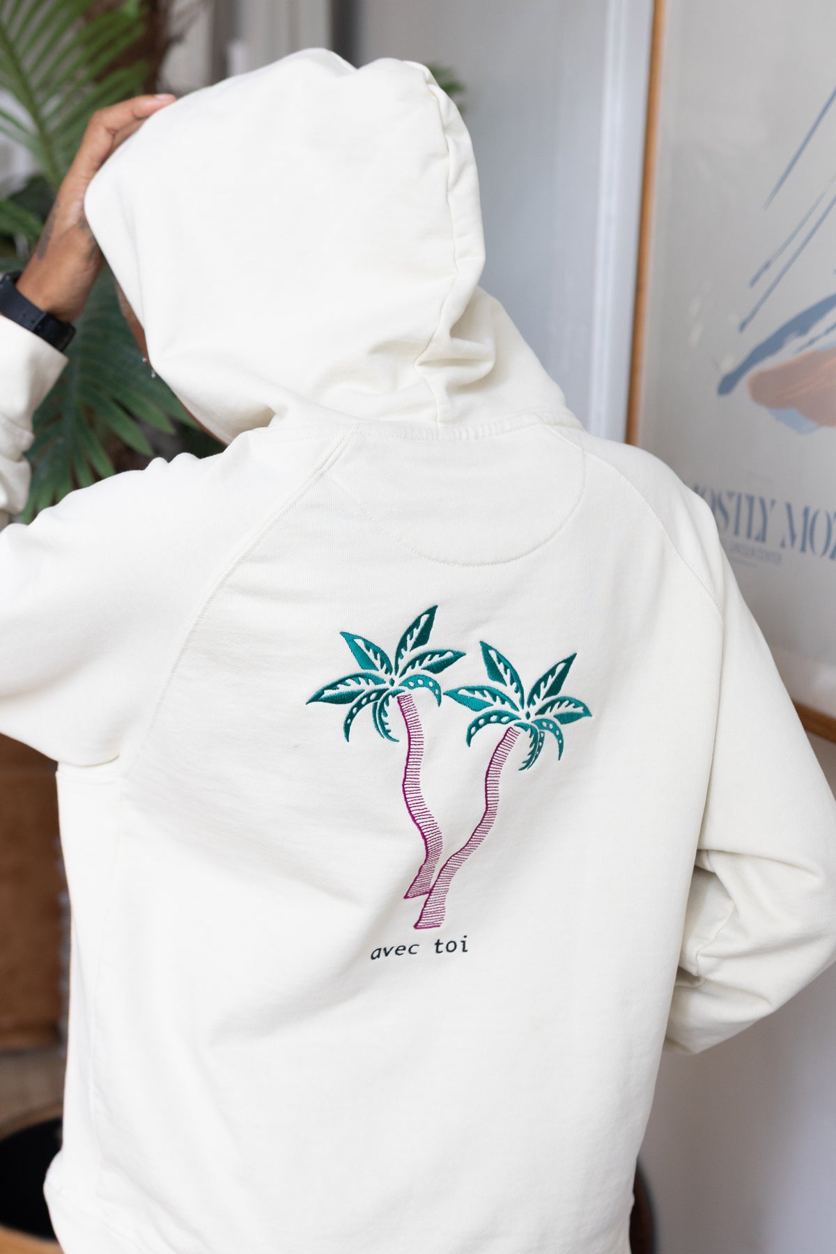 Back of the cream Avec Toi hoodie, with two palm trees embroidered in teal and magenta above the words “avec toi.”