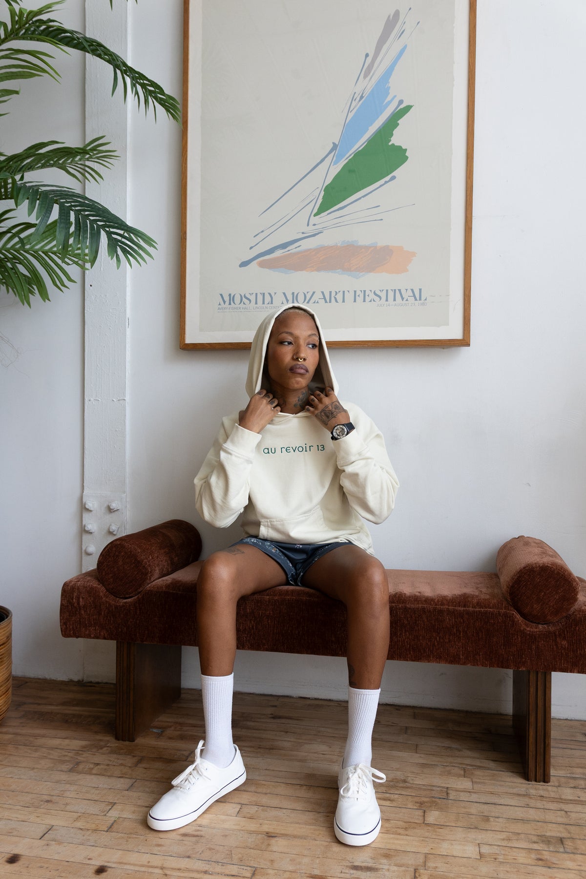 Woman in the cream Avec Toi hoodie with green Au Revoir 13 lettering, hood up, seated on a rust velvet bench.