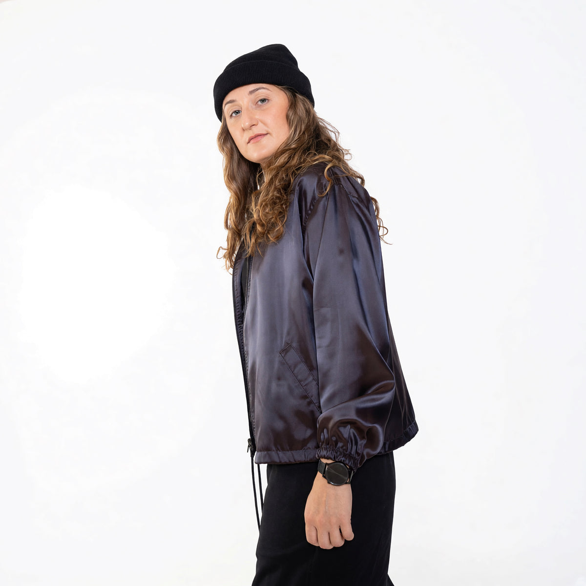 Side view of a woman in the Cher jacket in iridescent purple satin, zipped, with a black beanie and black pants.