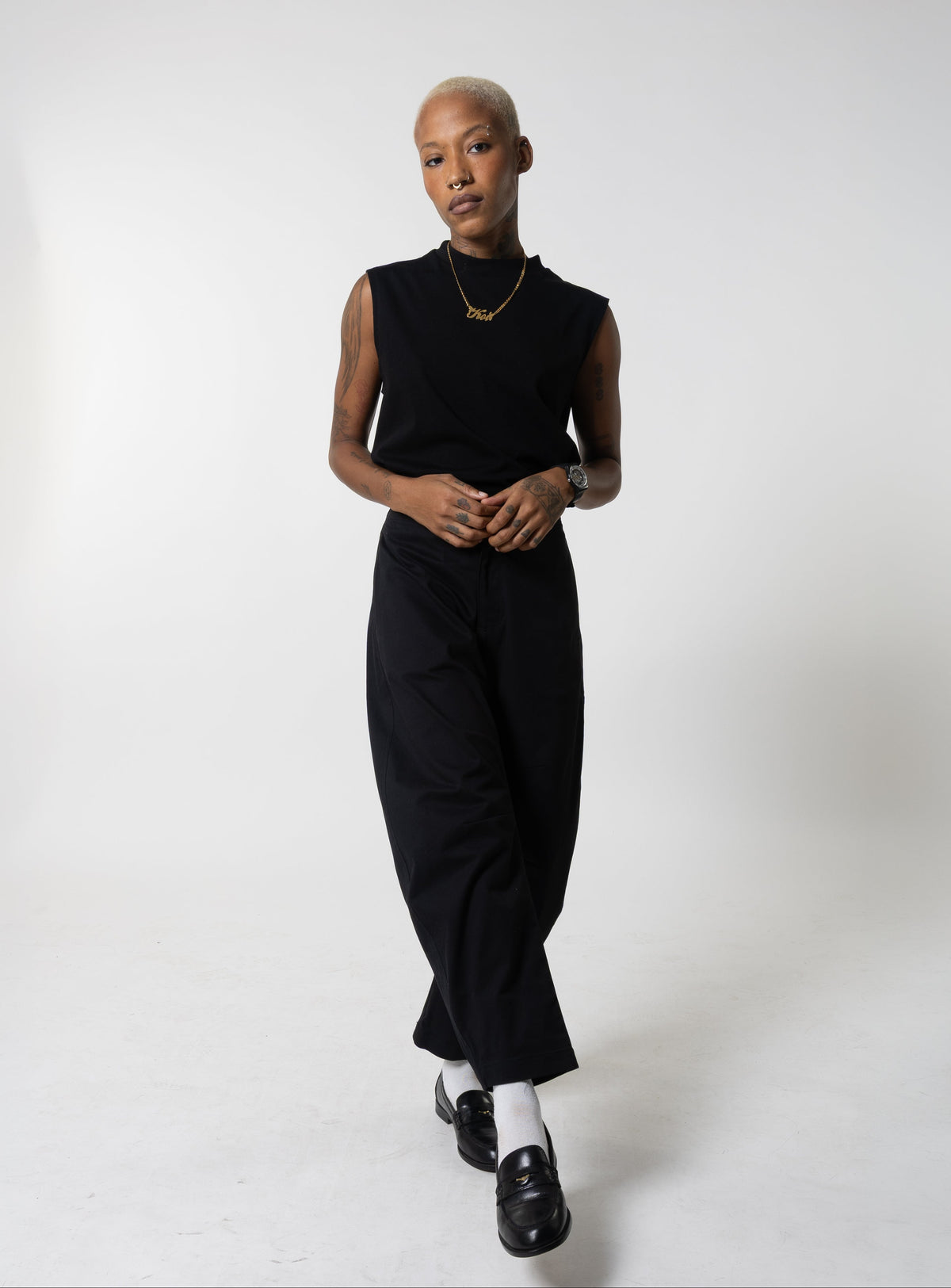 Woman in the black Elly tank and black wide-leg pants with a gold nameplate necklace and black loafers, mid-step.