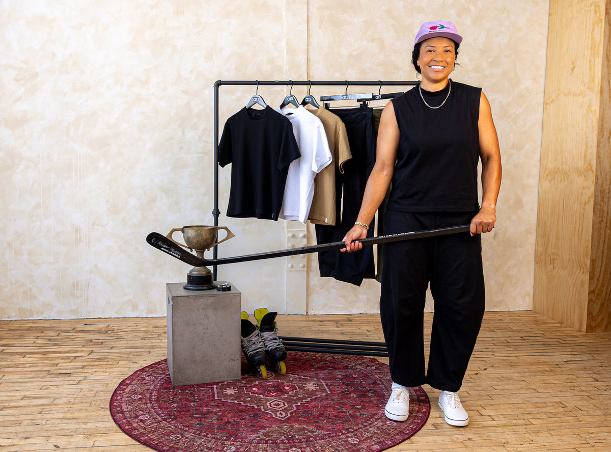 a13 founder in a black sleeveless mock-neck top and black barrel pants, holding a hockey stick beside a rack of a13 tees
