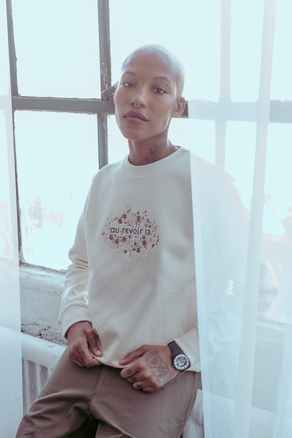 Woman in the cream Jame sweatshirt with the Au Revoir 13 logo framed in embroidered flowers, seated by a window.