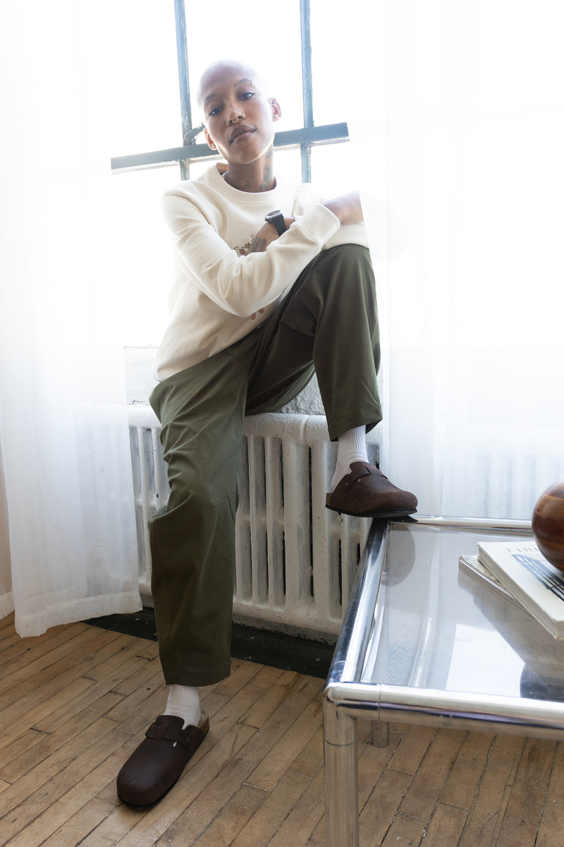 Woman in the cream Jame sweatshirt and olive green pants, seated on a radiator by a window with one clog on a glass table.