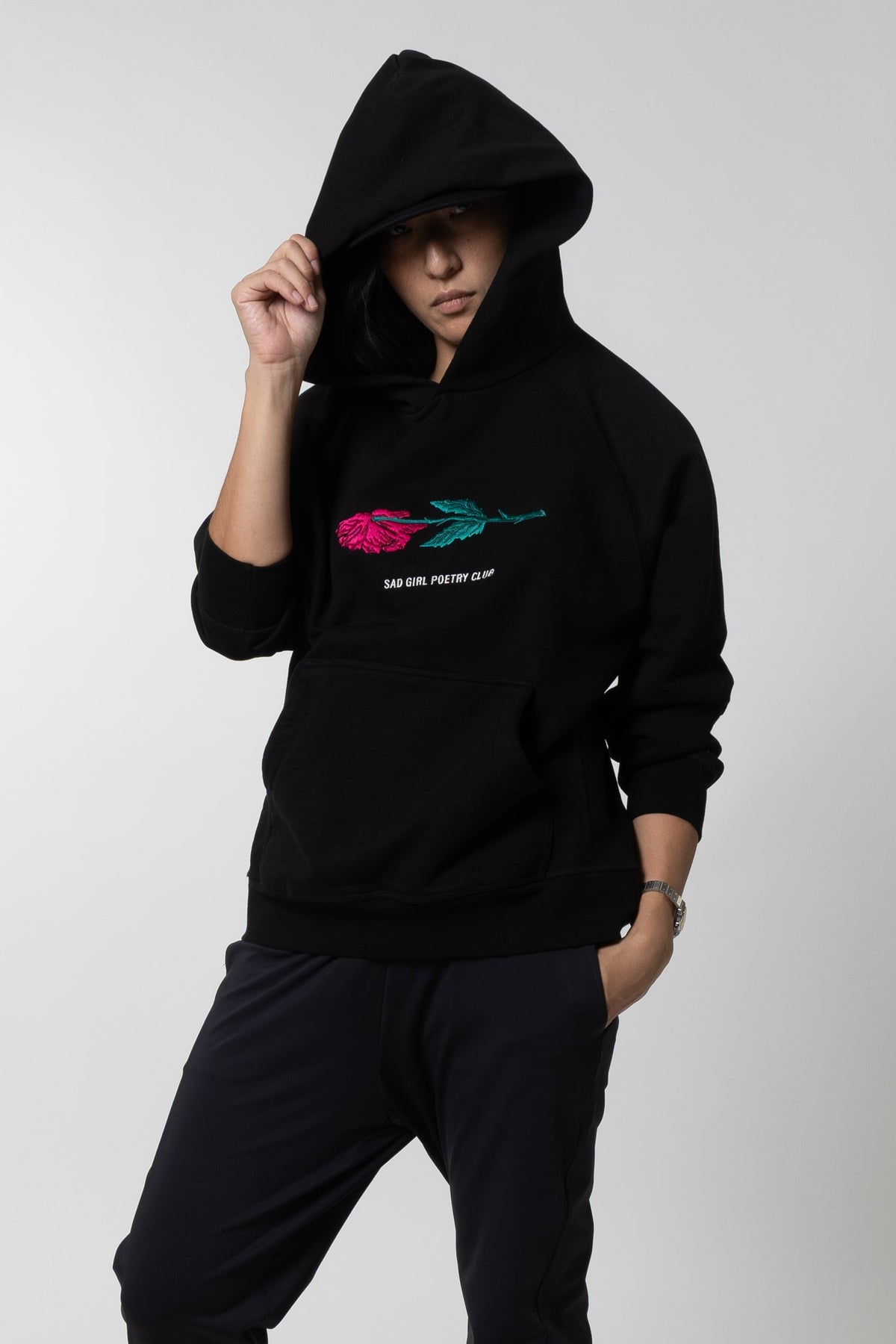 Woman in the black Sad Girl Poetry hoodie with the hood up, pulling it forward, showing the embroidered pink rose.