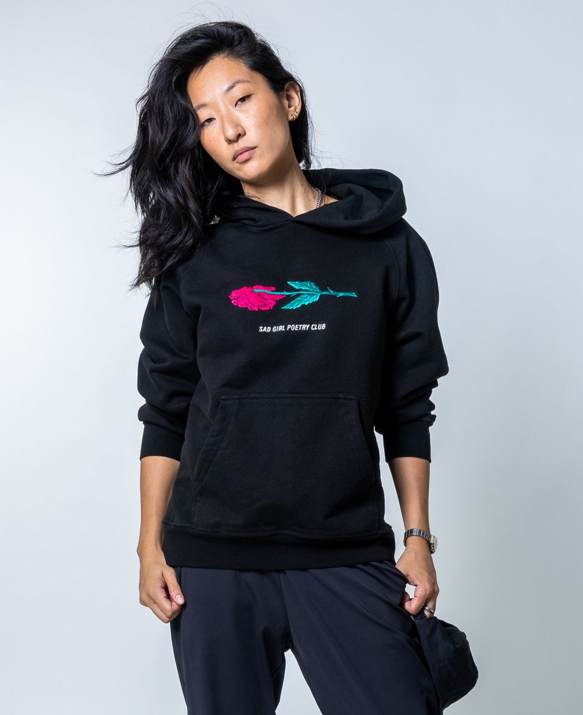 Woman in the black Sad Girl Poetry hoodie with an embroidered pink rose and teal stem above “Sad Girl Poetry Club.”