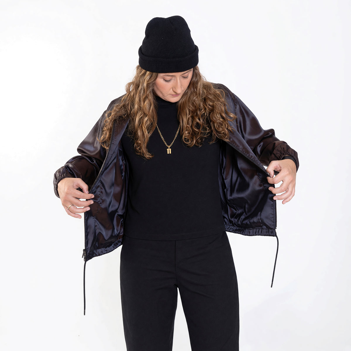 Woman opening the Cher jacket in iridescent purple satin, showing the drawstring hem, over an all-black outfit.