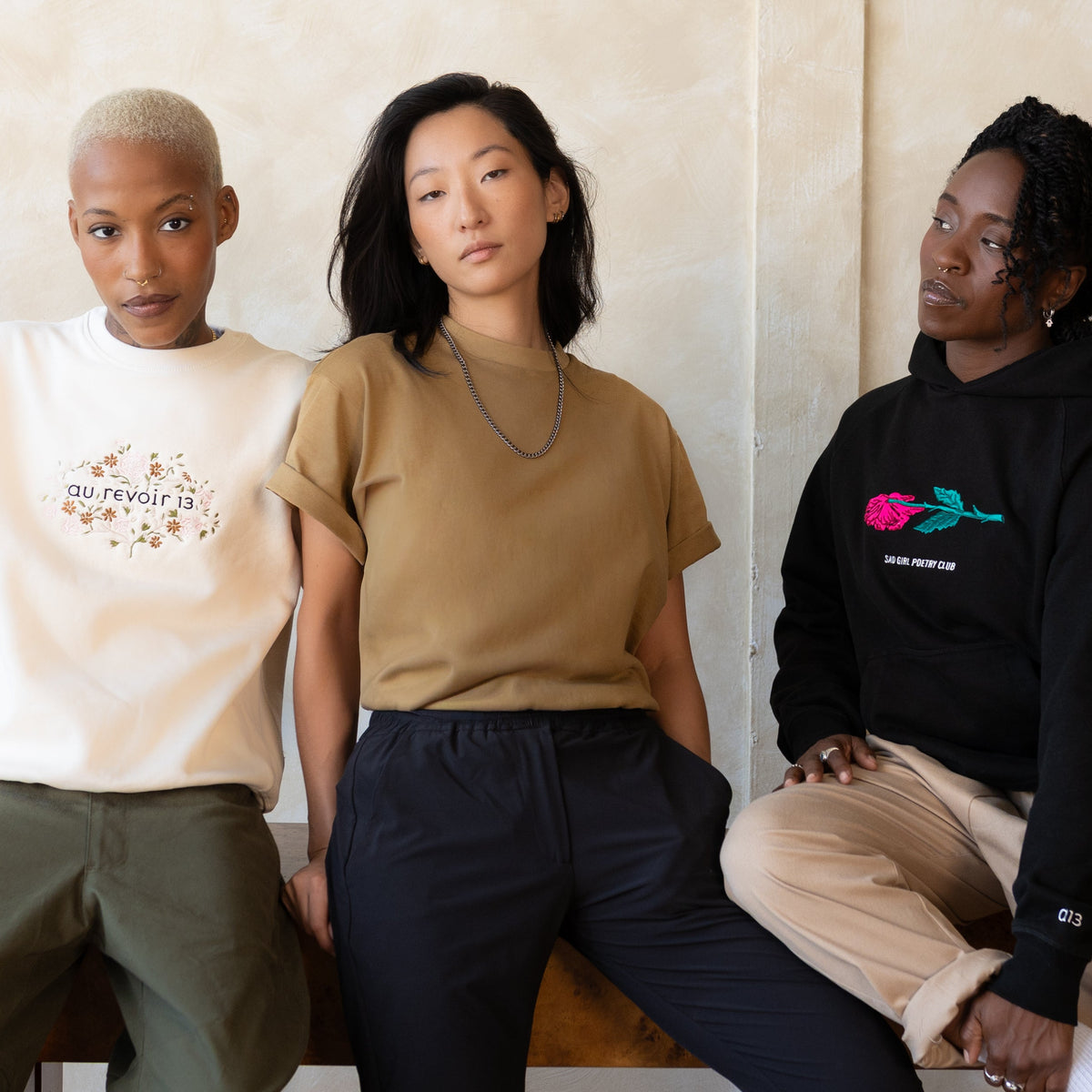 Three models in Collection 02: the cream embroidered Jame Crewneck, a tan boxy tee and the black Sad Girl Poetry Club hoodie